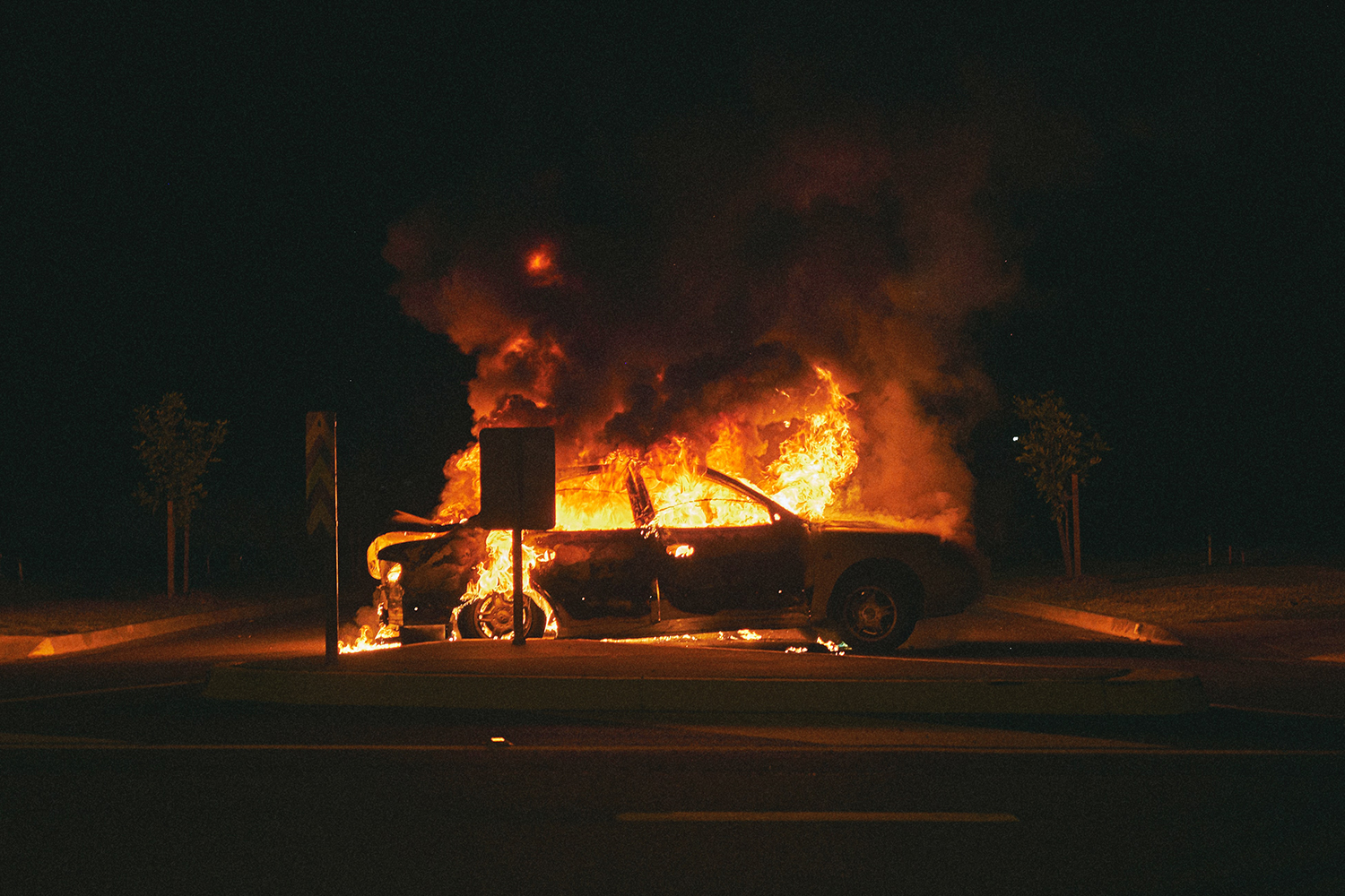 A four-door car on fire in a parking lot at night, with black smoke billowing up into the night sky