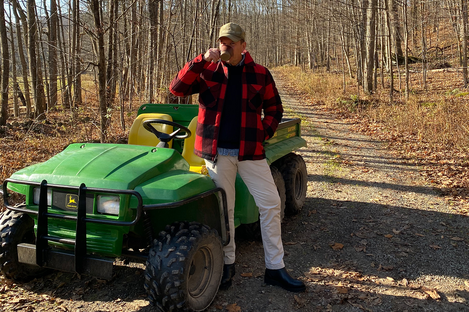 Photographer and magazine editor Matt Hranek sipping a cup of coffee in a buffalo plaid Woolrich jacket standing next to his John Deere Gator utility vehicle in the woods at his upstate New York estate