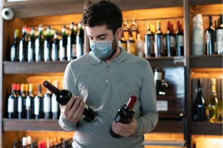 Young man at a wine cellar choosing between two wine bottles wearing a protective face mask. Covid-19 related supply issues are still affecting liquor stores in some states.