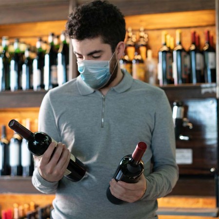 Young man at a wine cellar choosing between two wine bottles wearing a protective face mask. Covid-19 related supply issues are still affecting liquor stores in some states.