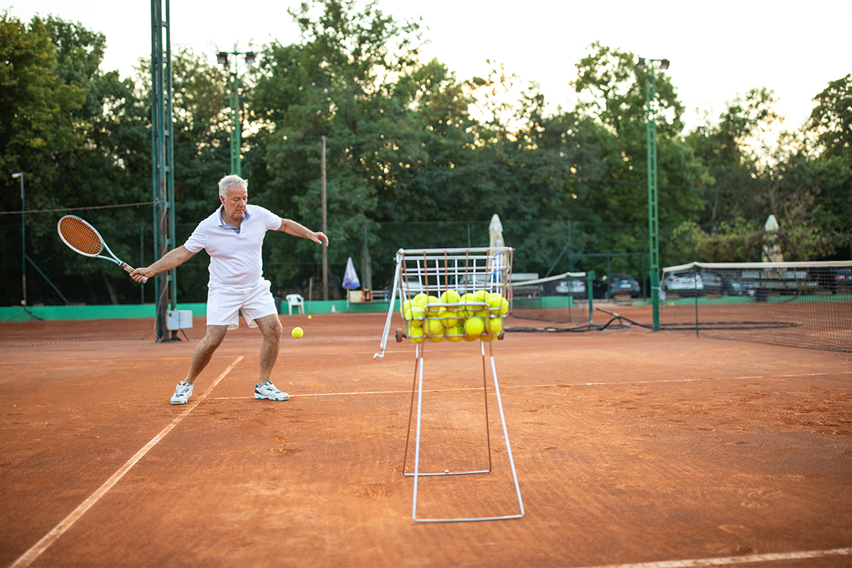 An old man playing tennis on clay courts with a basket of tennis balls in the foreground