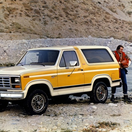 A white and yellow 1982 Ford Bronco SUV sitting among rocks and a river next to two outdoorsmen in a retro photo