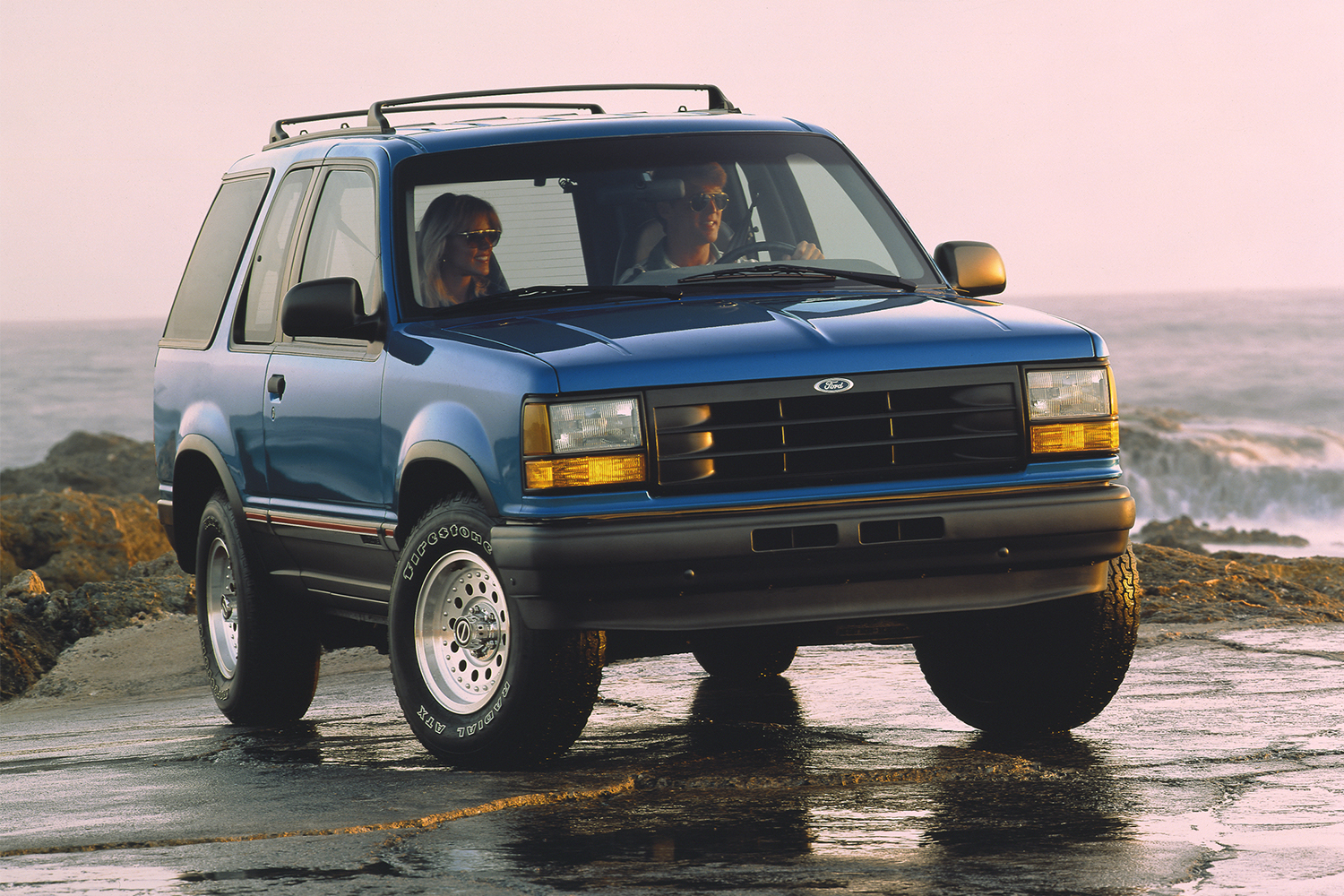 A man driving a 1992 blue Ford Explorer SUV near the ocean in a vintage photo