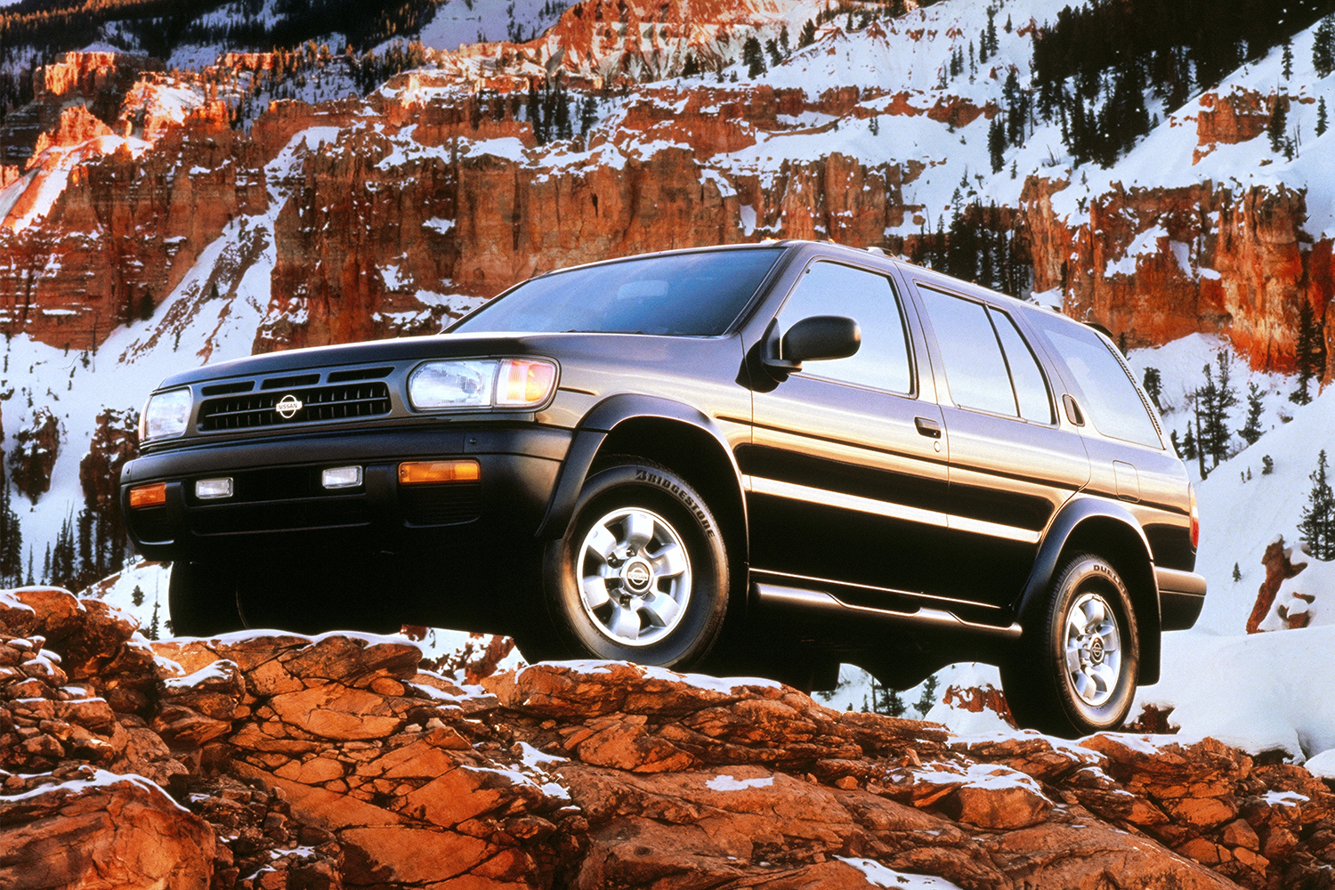 A 1997 Nissan Pathfinder SUV perched on a mountain with snow covered rock in the background in a vintage photo