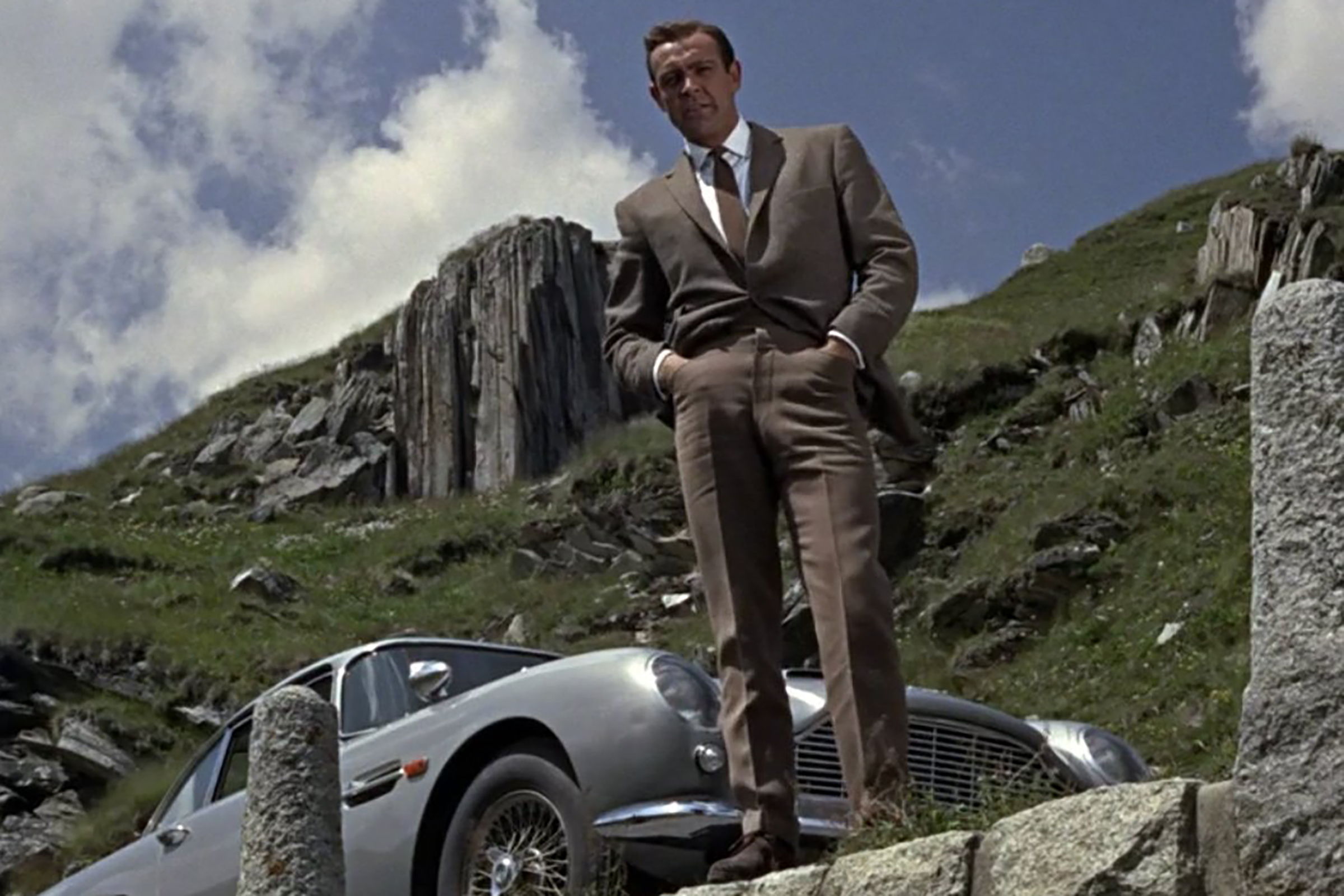 Connery wears a brown knit tie with a white shirt and tan trousers with a perfect half break in Goldfinger.