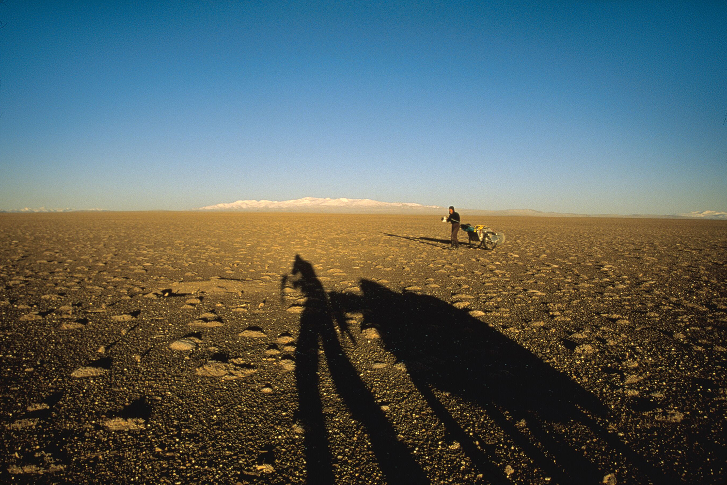 Rick Ridgeway pushing a cart across the Chang Tang Plateau in Tibet in 2002 with the shadow of Galen Rowell in the forground and an uninhabited expanse behind