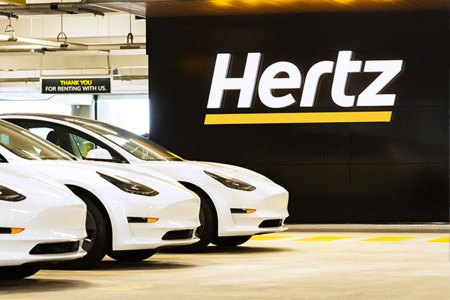 Three white Tesla Model 3 electric vehicles sitting next to a Hertz sign. The rental car company ordered 100,000 EVs from Elon Musk's car company.