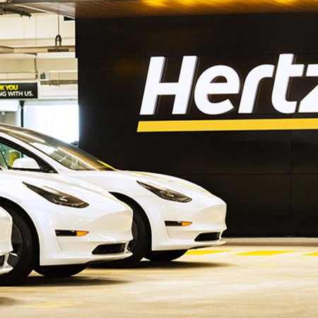 Three white Tesla Model 3 electric vehicles sitting next to a Hertz sign. The rental car company ordered 100,000 EVs from Elon Musk's car company.