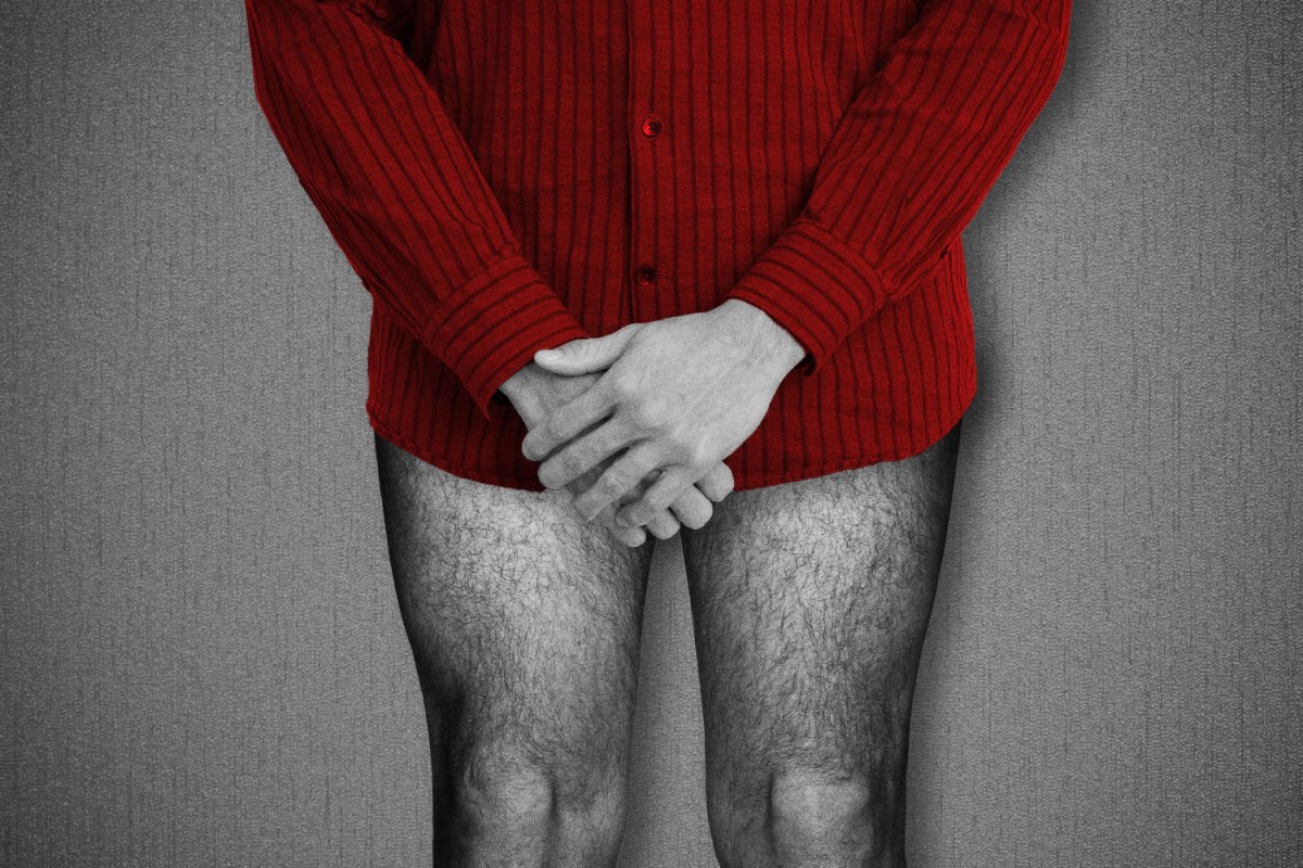 Black and white photo of a man wearing a red shirt and no pants with his hands clasped over his crotch
