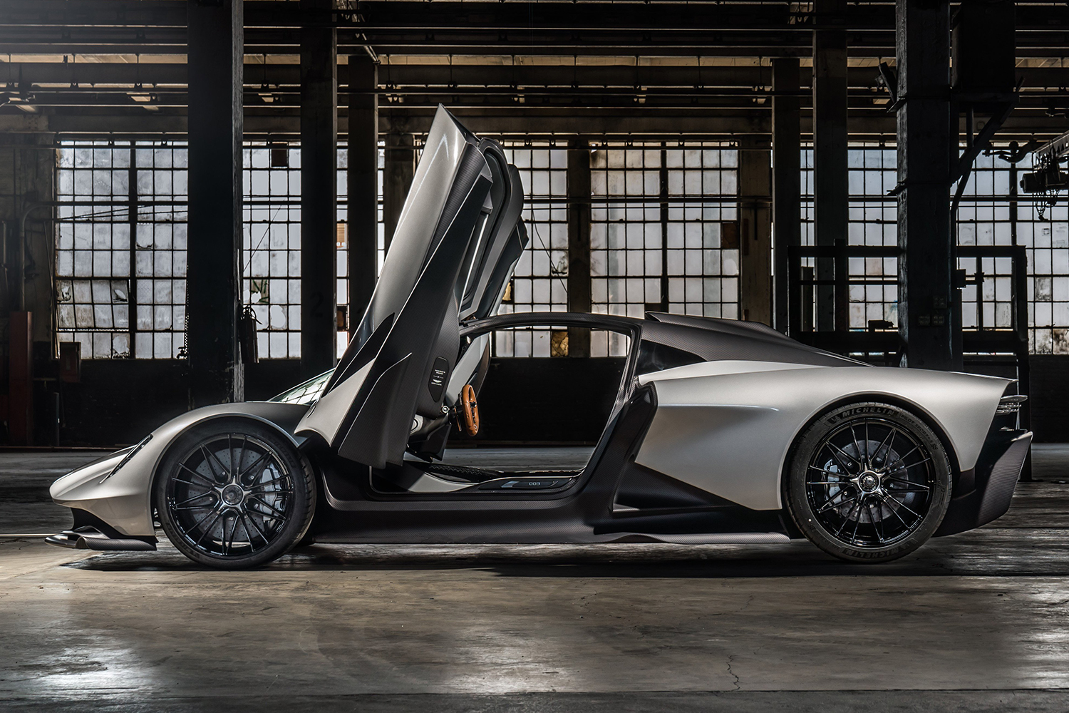 A side profile of the original Aston Martin Valhalla hypercar with the wing doors open so you can see through the car to the warehouse windows behind it