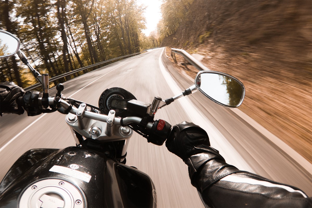 The handlebars and side mirror of a motorcycle as it speeds down a winding road in the woods. A motorcycle just a record for the Cannonball Run, an illegal nationwide road race.