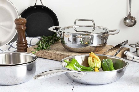 French Cristel stainless steel skillets and nonstick pans on a table with a cutting board and some assorted vegetables