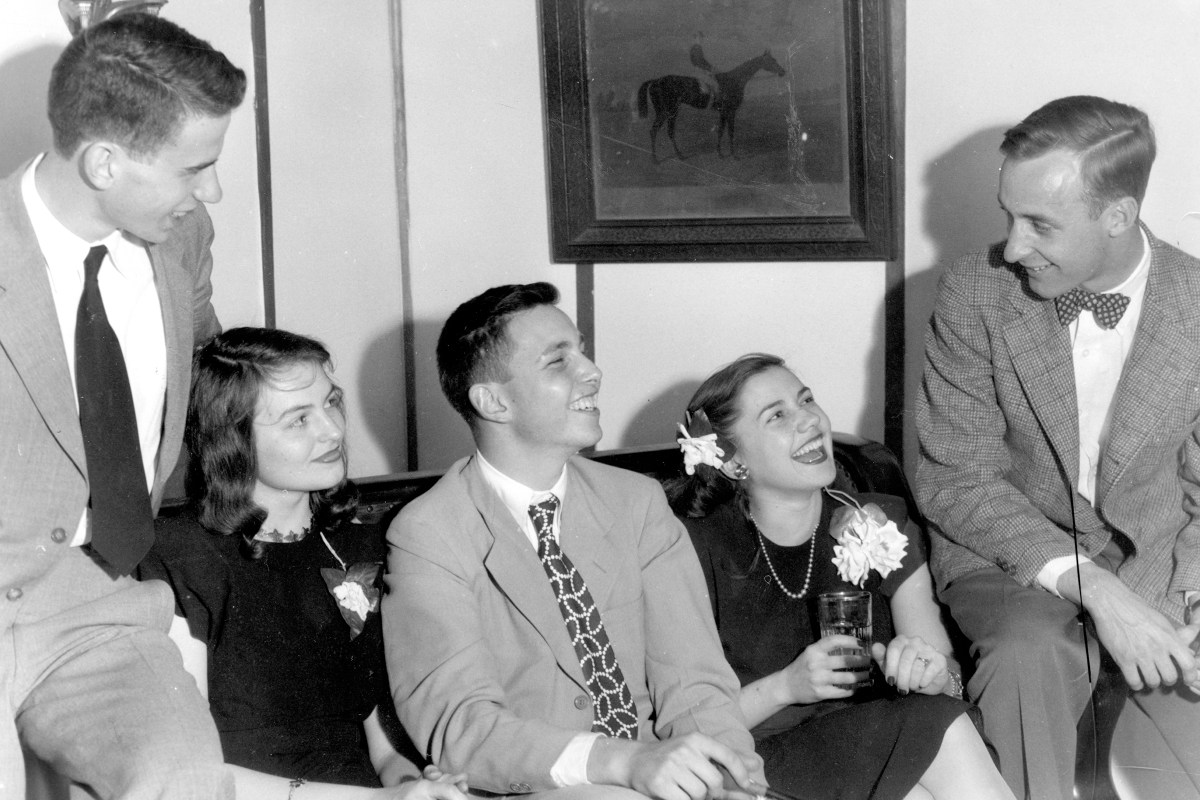 At a party hosted by the Delta Phi fraternity at Johns Hopkins University, brothers lounge on a sofa in a group, wearing suits next to their girlfriends in dresses and corsages, everyone smiling and drinking beverages from glasses, in a fraternity house right off campus, Baltimore, Maryland, 1947.