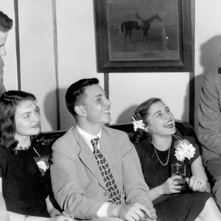 At a party hosted by the Delta Phi fraternity at Johns Hopkins University, brothers lounge on a sofa in a group, wearing suits next to their girlfriends in dresses and corsages, everyone smiling and drinking beverages from glasses, in a fraternity house right off campus, Baltimore, Maryland, 1947.