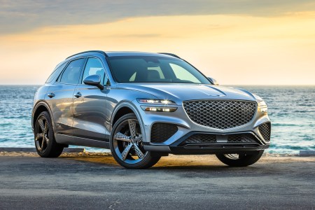 The Genesis GV70 SUV with the headlights on sitting still with a beach and sunset in the background
