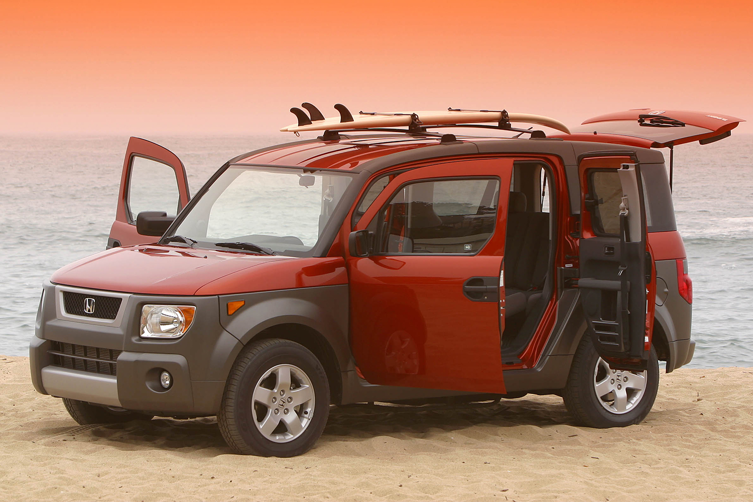An orange and brown Honda Element with all the doors open and a surfboard on top sitting on the beach at sunset