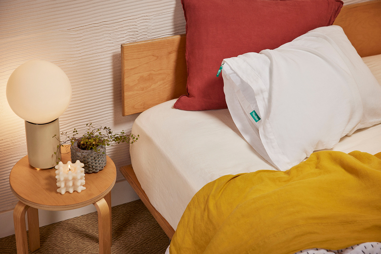 A Marlow pillow from Brooklinen's new sub-brand sitting on a bed with yellow sheets and a red pillow next to a nightstand