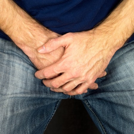 Close up photo shows a man holding his hands folded over his groin while wearing blue jeans