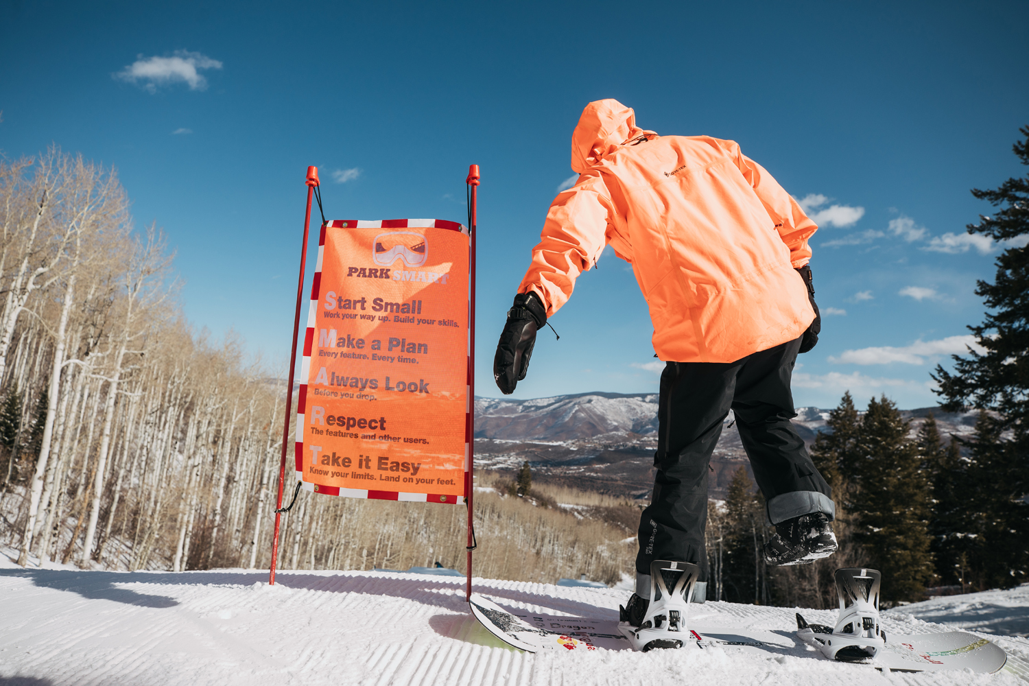 Pro snowboarder Brock Crouch riding Burton Step On strapless bindings. Here he's pictured in an orange winter jacket on the top of a snowy mountain in Aspen, Colorado next to an orange sign.