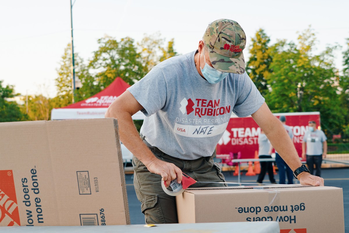 A man with a camo Team Rubicon hat and grey Team Rubicon T-shirt tapes a cardboard box with donations for Afghan families resettling in the U.S.