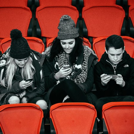 Three young people in the stands at a professional soccer game browse their smarthpones