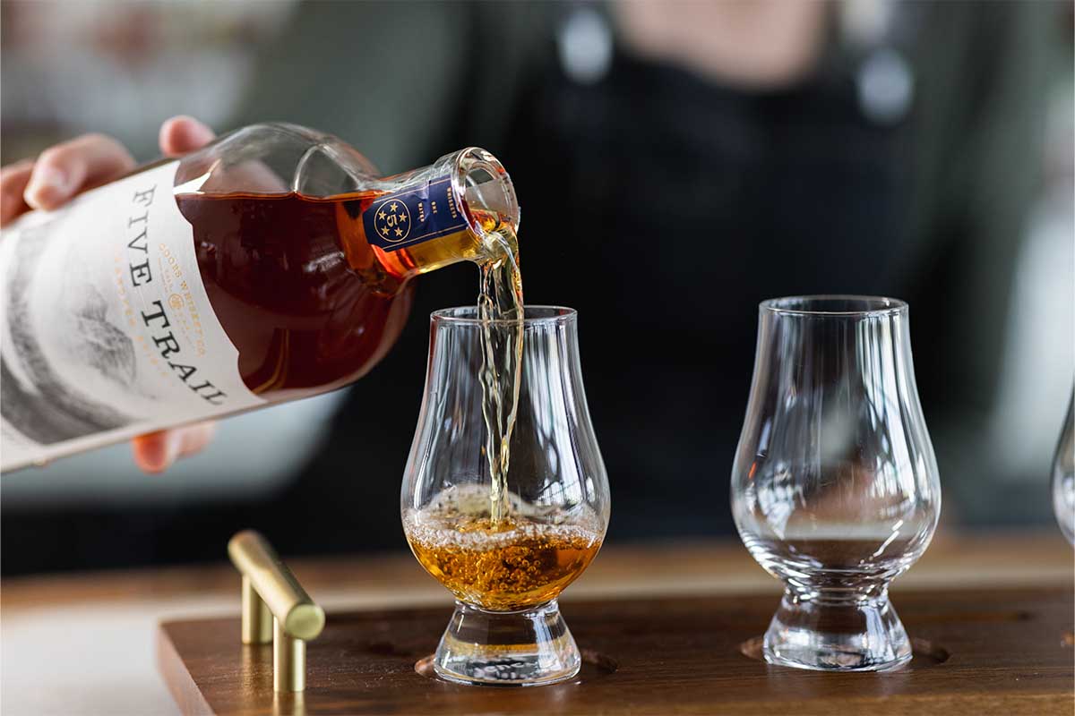 A hand pouring Five Trail American Whiskey into two tasting glasses. Five Trail is the first release from Coors Whiskey.