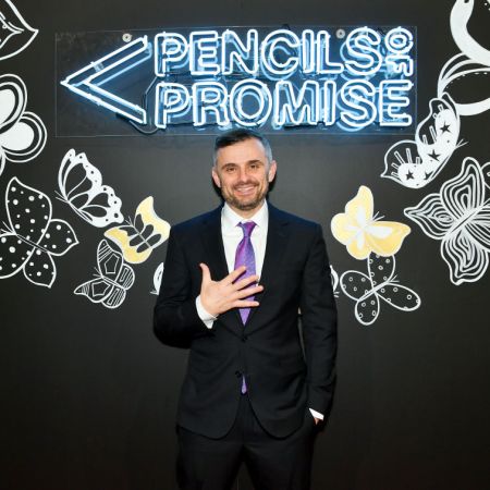 Entrepreneur Gary Vaynerchuk in a black suit with a white shirt and purple tie standing in front of a neon sign that says "Pencils of Promise"
