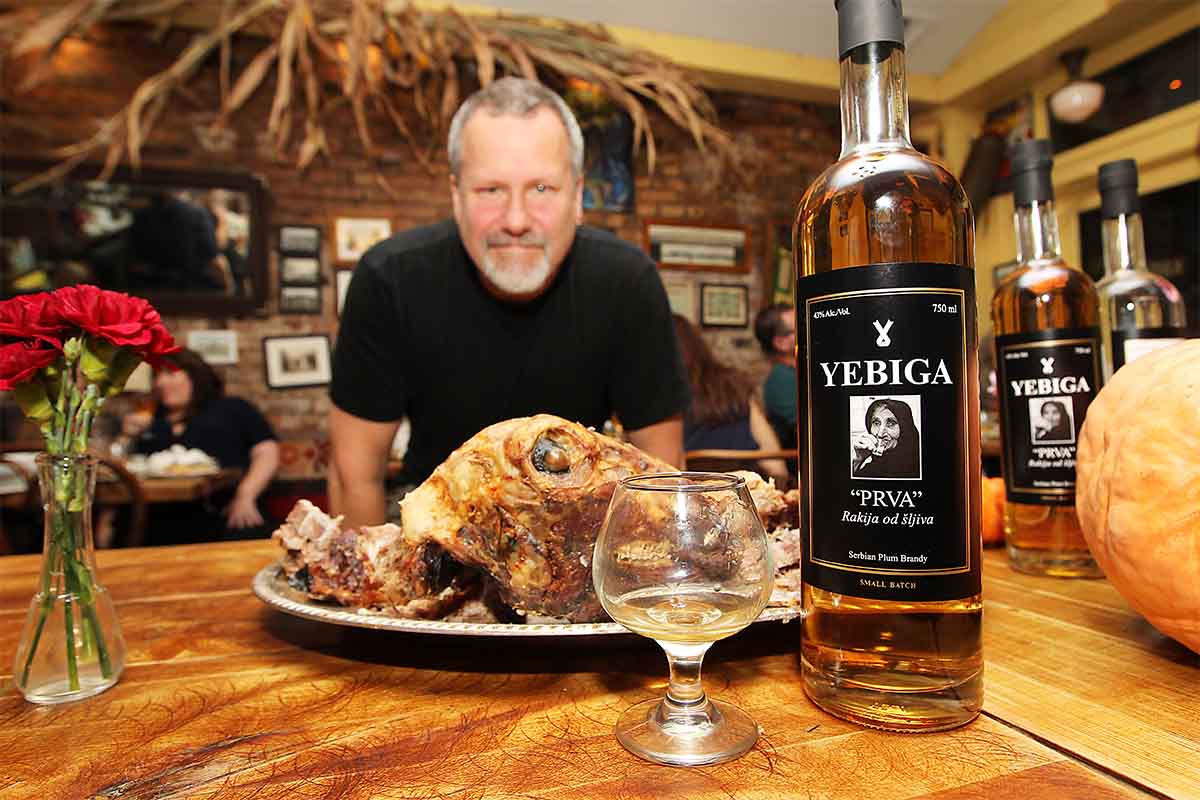 Bill Gould posing with a bottle of Yebiga rakija in front of a plate of meat. The musician is importing rakija to the U.S. from Serbia.