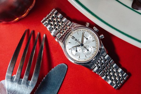 A vintage Omega Seamaster Chronograph sitting on a red tablecloth on a dining table next to a fork, knife and plate. The vintage watch is currently on sale through J.Crew.