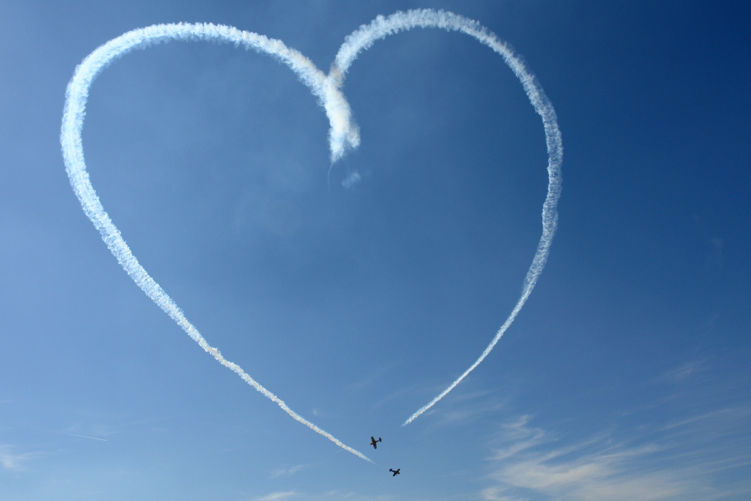 Two airplanes leaving a contrail heart in an almost clear sky. A new plane service will allow couples to enjoy the mile high experience without a destination