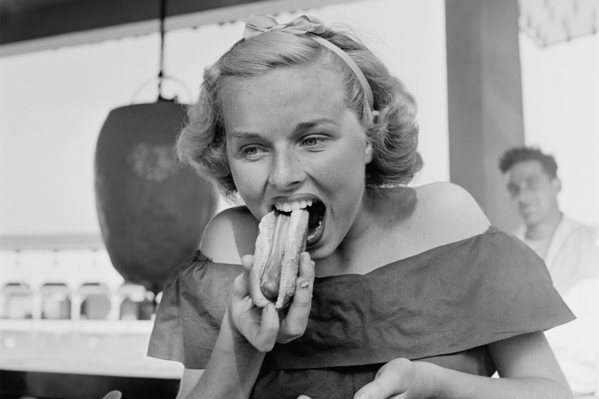 Black and white photo shows a young woman eating a hot dog at Palisades Park, New Jersey, 1949.