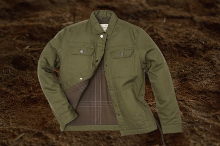 Tentree's Treeline Canvas Unisex Jacket in olive green on a dirt background. The outerwear is supposedly 99% compostable.