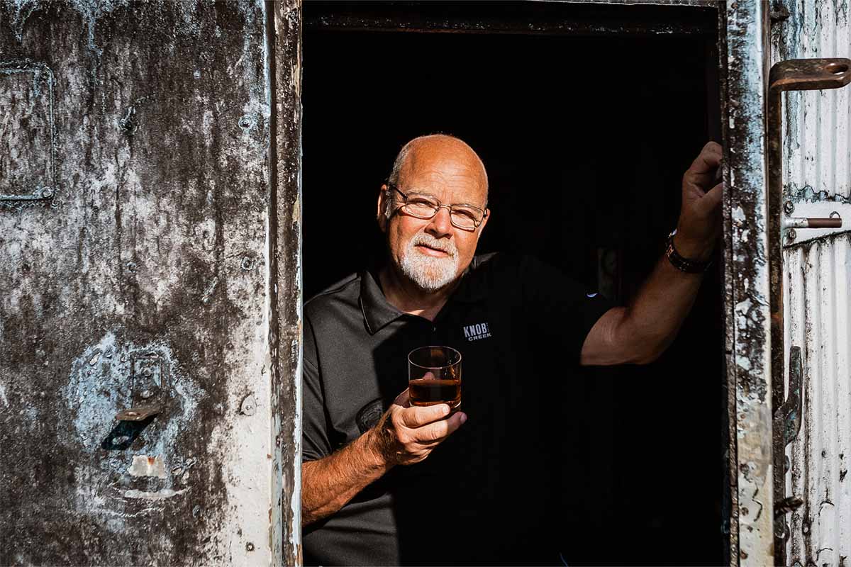 Master Distiller Fred Noe holding a glass of whiskey and leaning against a door