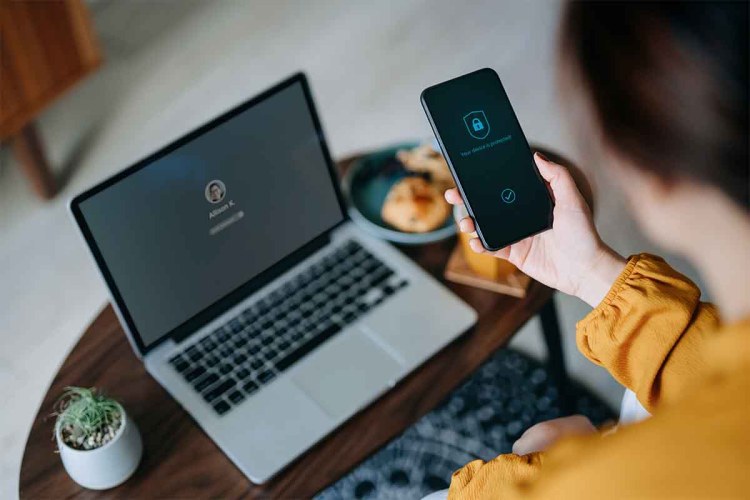 stock photo of Young Asian woman logging in to her laptop and holding smartphone on hand with a security key lock icon on the screen, sitting in the living room at cozy home. Password security for corporations was recently called out by Dashlane.