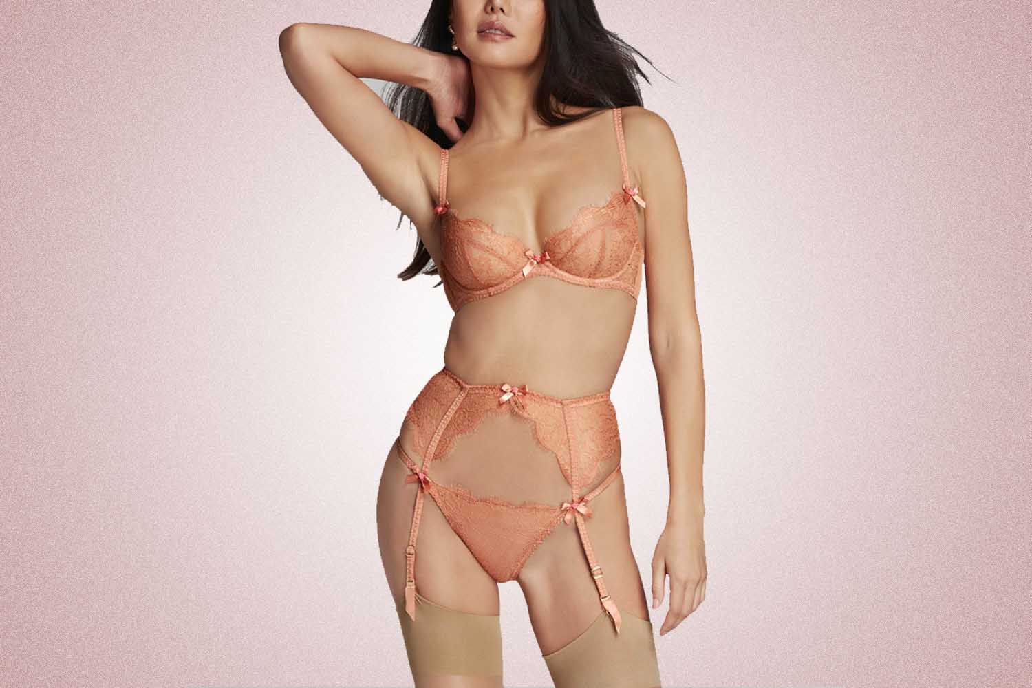 A woman wearing a light pink, lacy lingerie set that includes a bra, suspender and thong, a perfect Valentine's Day gift, on a pink background. 