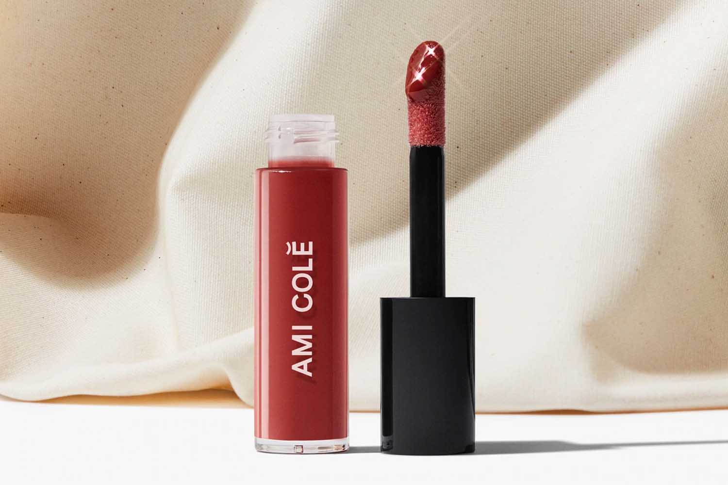 A small tube of lip oil next to a black lip gloss applicator from Ami Cole, a perfect Valentine’s Day gift for 2022, on a tan background. 