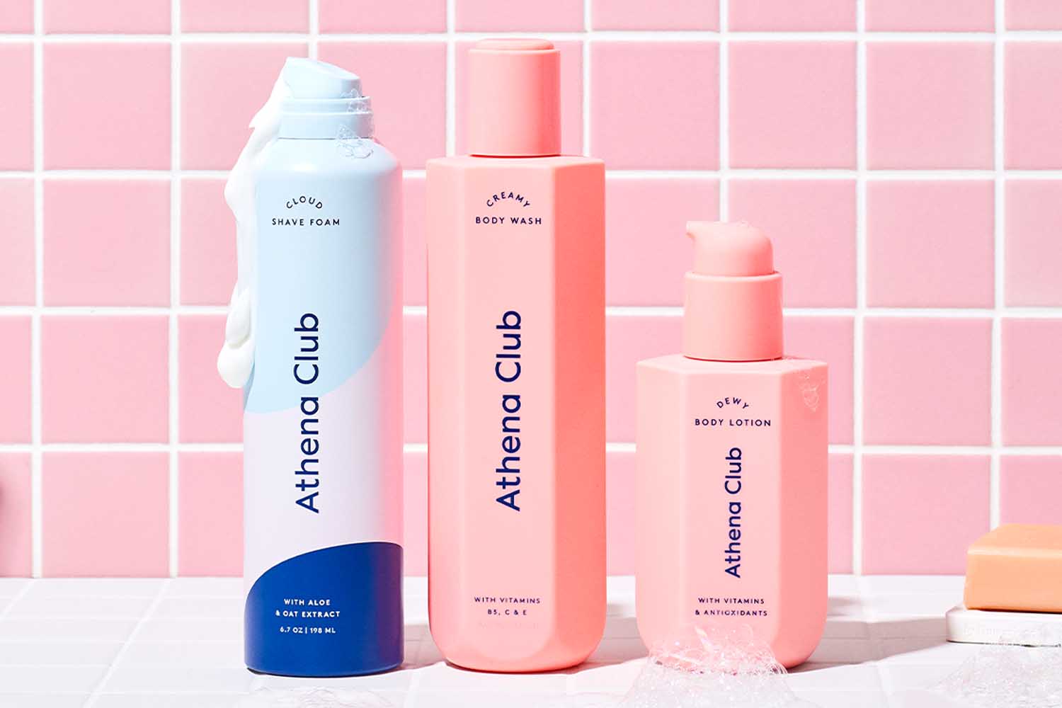 A blue can of shaving cream, pink bottle of body wash and a pink bottle of body lotion from the Athena Club on a pink tile background, a perfect Valentine’s Day gift for 2022.