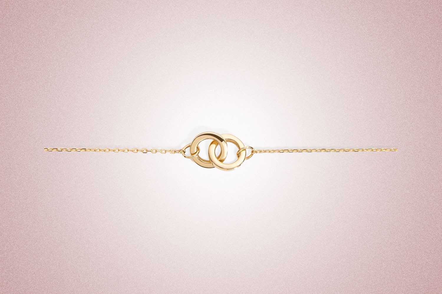 A gold bracelet with two interlocking loops at the center from Aurate, a perfect Valentine’s Day gift for 2022, on a pink background.