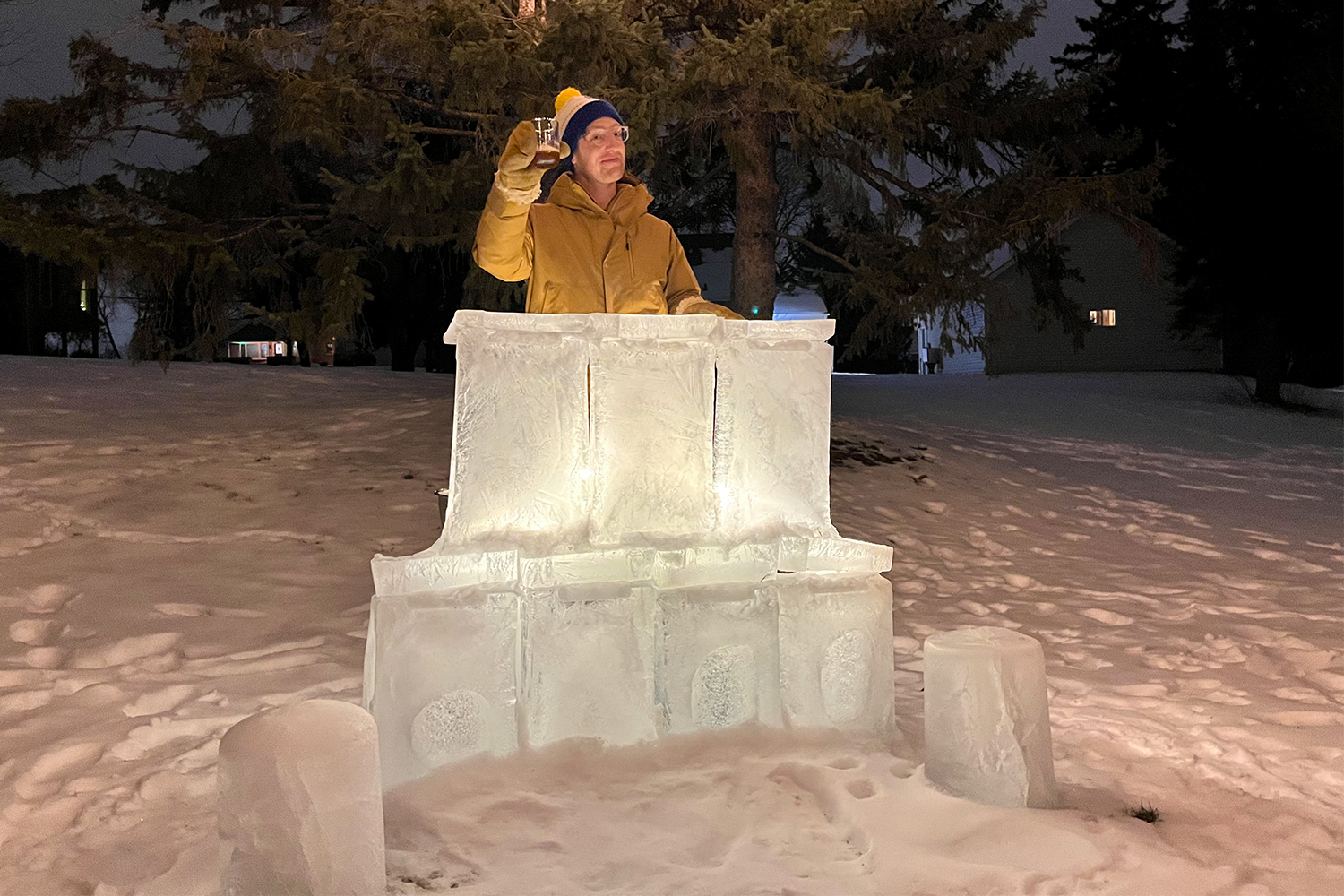 A man in a winter jacket, mittens and beanie holding up a cocktail from a homemade ice bar. In this article, we talk about how to build your own ice bar.