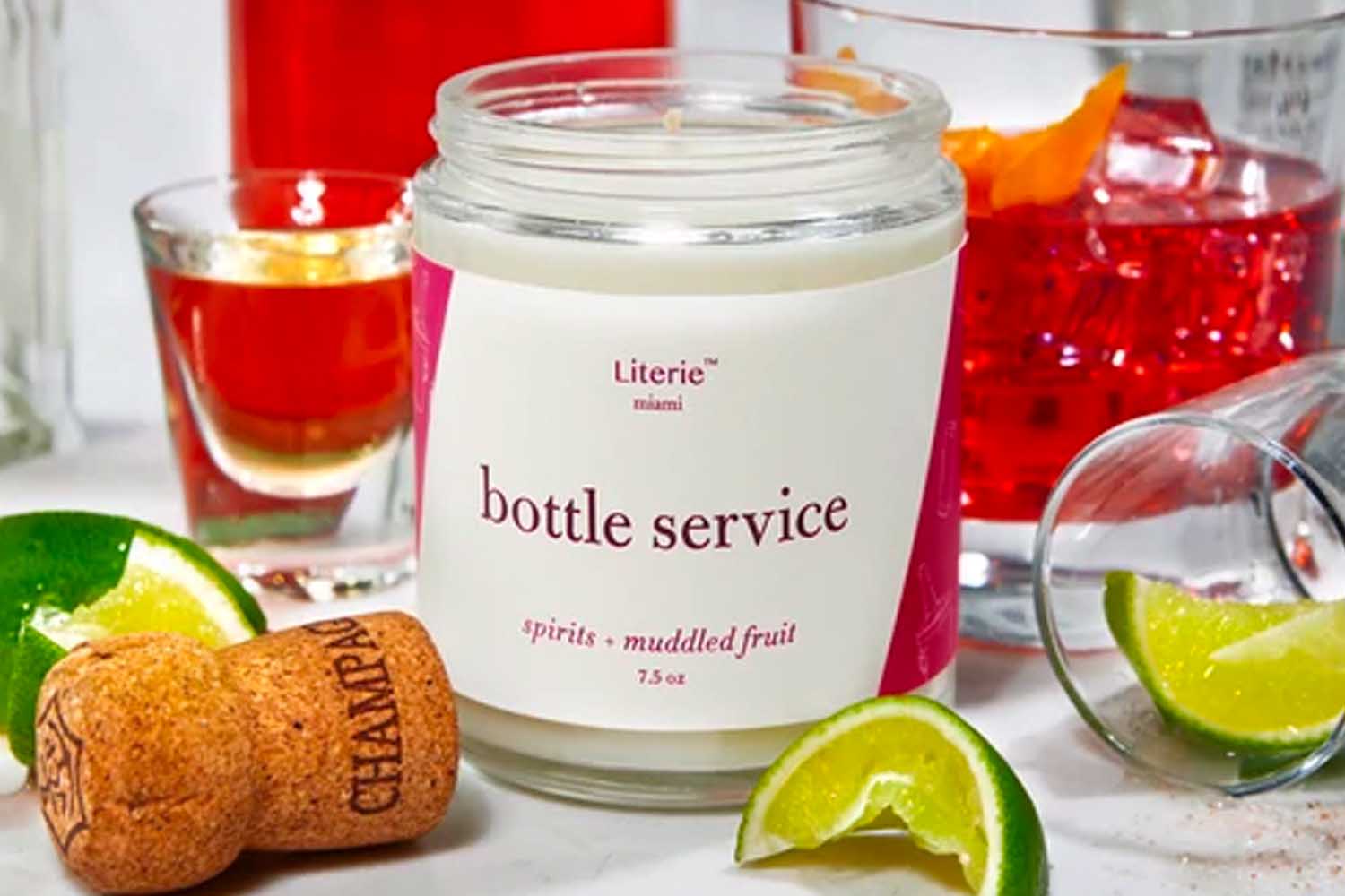 A white candle with a white and purple label that reads "bottle service, spirits + muddled fruit" surrounded by cocktails, limes and a champagne cork from Literie, a perfect Valentine’s Day gift for 2022.
