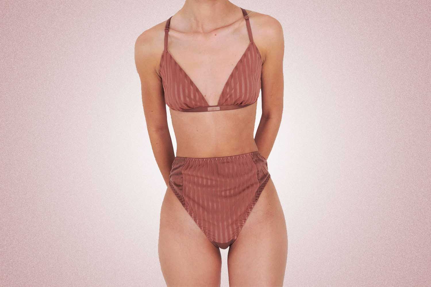 A woman wearing a tan pinstripe set with high waisted briefs and a triangle bra from Lunya, a perfect Valentine's Day gift, on a pink background. 