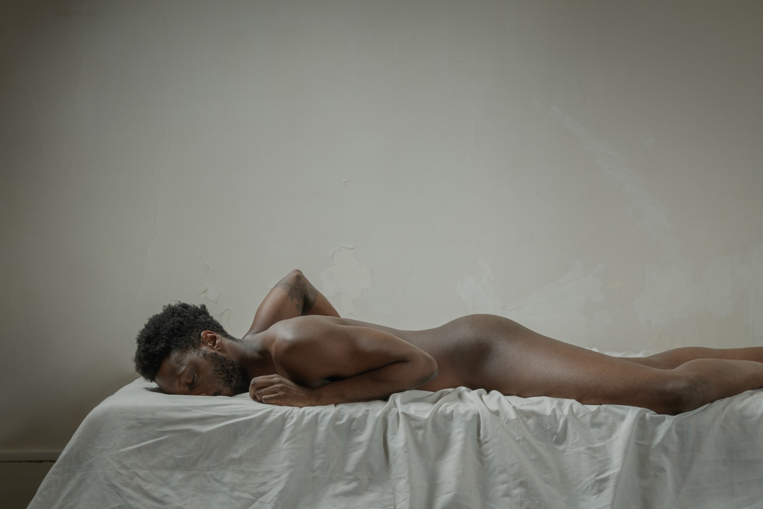 A man lies face down on a white bed, posing for Laura Stevens' series of male nudes, "Him"