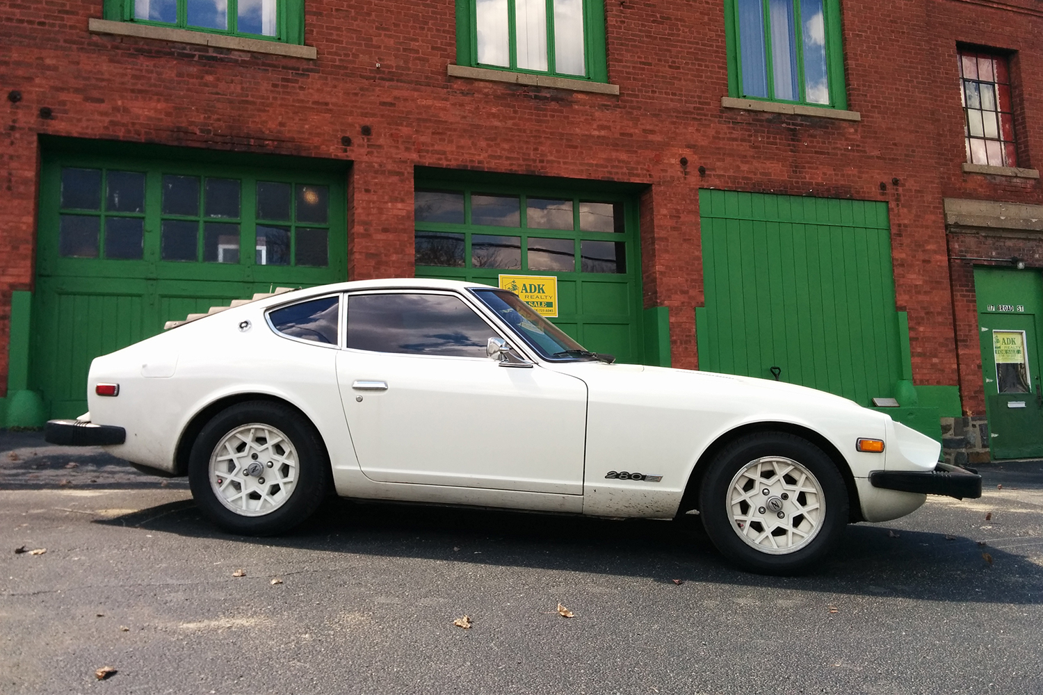 A side profile of a white 1978 Datsun 280Z with snowflake wheels sitting in front of a brick building with green doors. Here's what you need to know before you buy one.