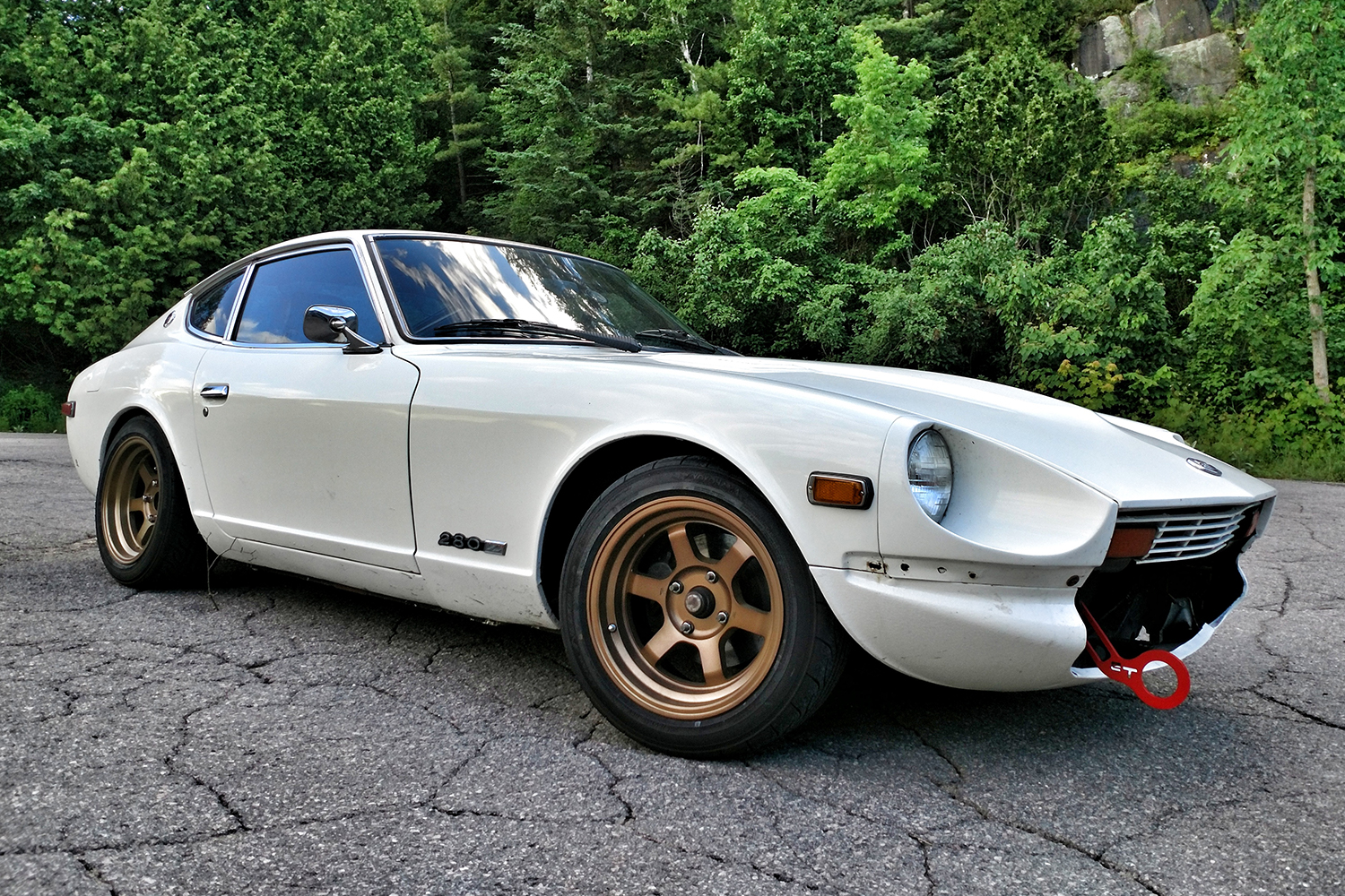 A white 1978 Datsun 280Z sports car sitting on asphalt in front of green trees. In this story, the Datsun owner talks about what it's like to own the '70s car.