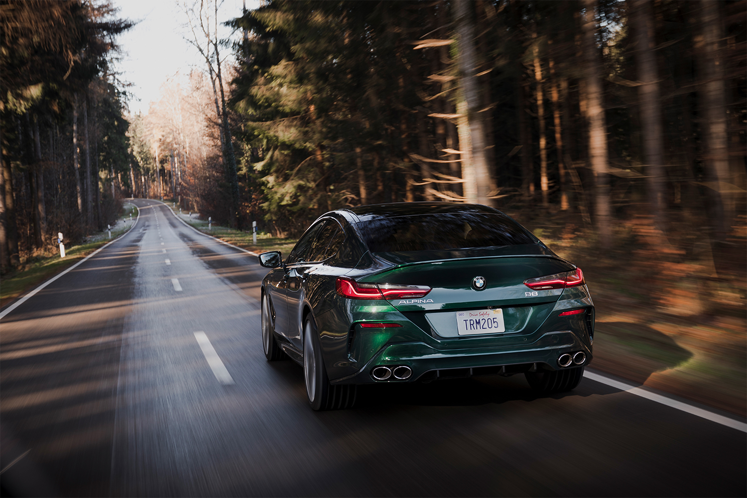 The 2022 BMW Alpina B8 Gran Coupe in green shown from the rear while it drives down a road between lines of trees. In our review of the luxury car, we found it excels in comfort and speed.