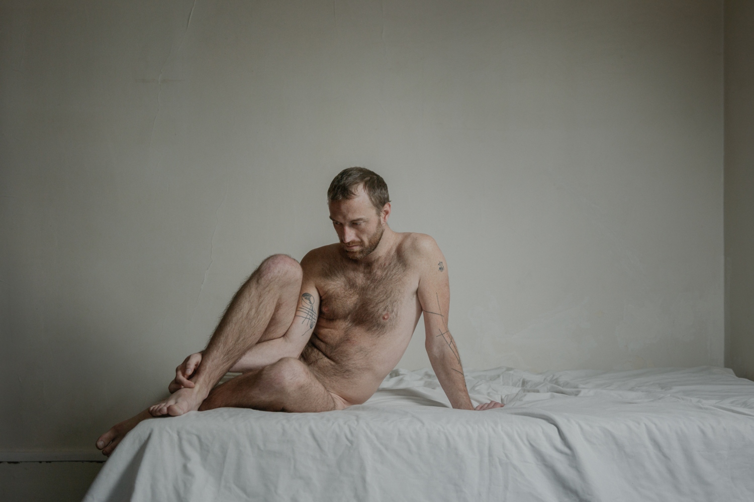 A nude man poses sitting on a white bed for a photo by Laura Stevens