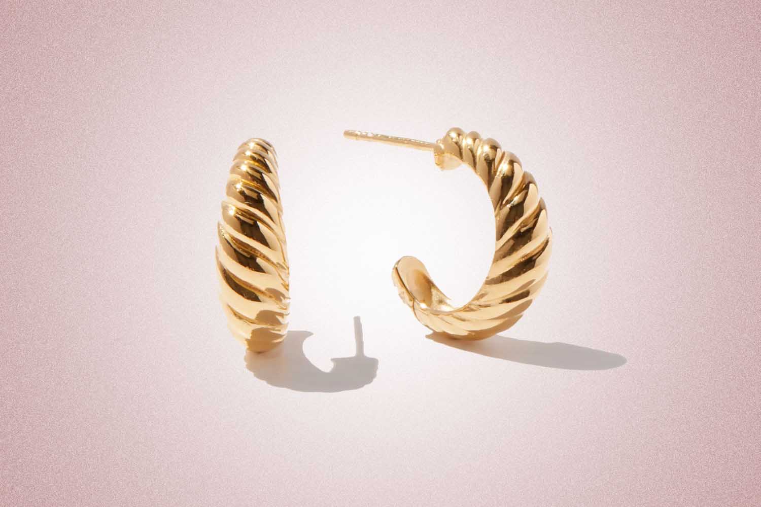 Gold croissant shaped hoop earrings, a perfect valentine's day gift, on a pink background.