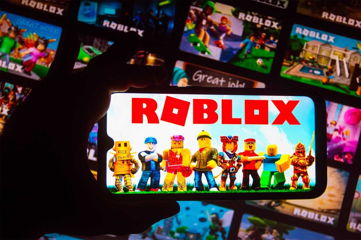 Phone with Roblox characters held in front of a screen of Roblox games.