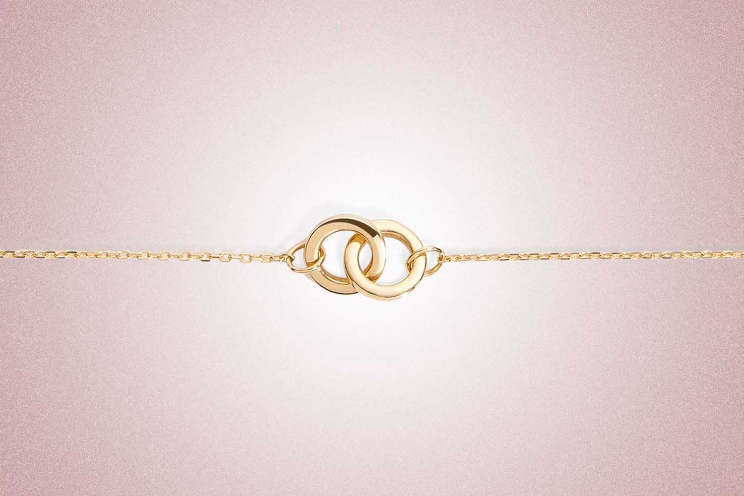 A gold chain bracelet with two interlocking loops at the center from Aurate, a perfect valentine's day gift, on a pink background