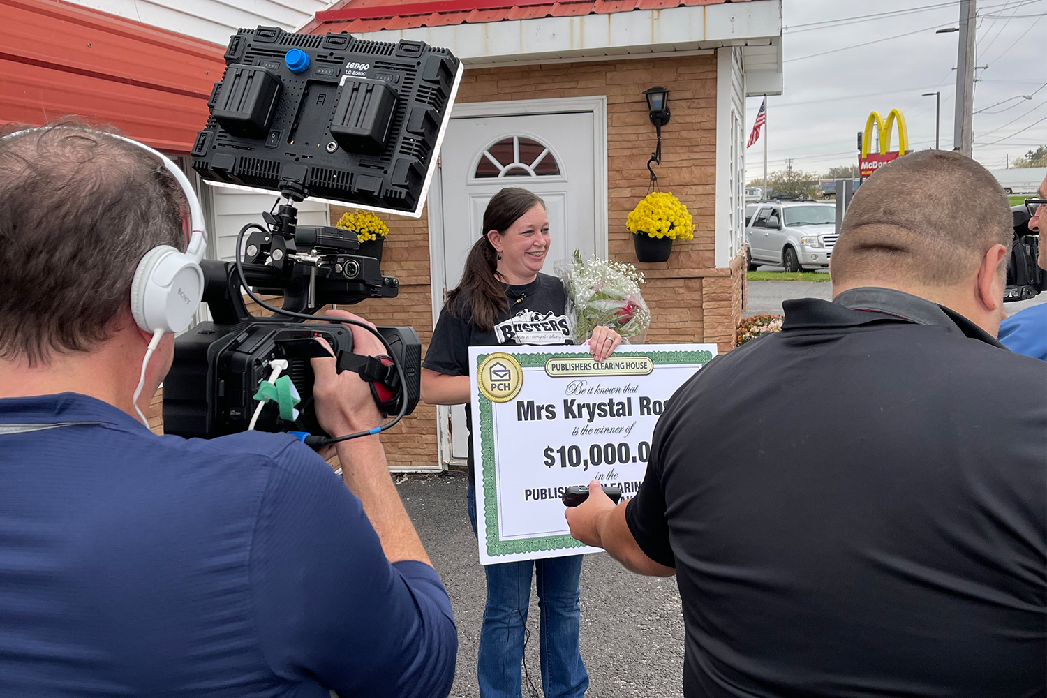 Krystal Rose, who worked at Busters in Ogdensburg, New York, standing in front of cameras and reports while holding a ceremonial check for $10,000 from Publishers Clearing House, presented to her by Prize Patrol member Howie Guja.